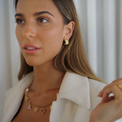 A close-up lifestyle shot of a woman wearing a gold charm necklace with small heart and flower charms, a gold hoop earring, and a gold ring, paired with a textured white jacket.