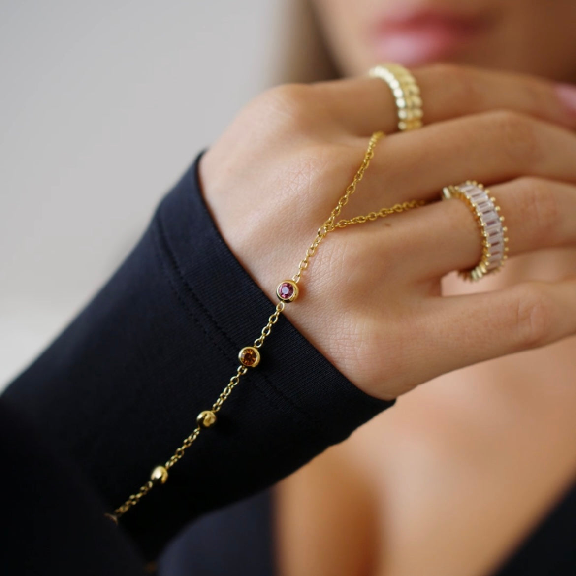 A close-up lifestyle shot of a delicate gold hand chain bracelet with 5 small cz  colorful gemstone, worn on a model's wrist alongside gold rings.