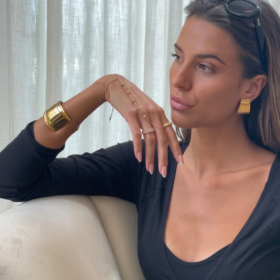 A woman wearing gold jewelry, including a chunky gold cuff bracelet, a delicate gold chain bracelet, rings, and textured gold earrings, posing indoors in front of white curtains.