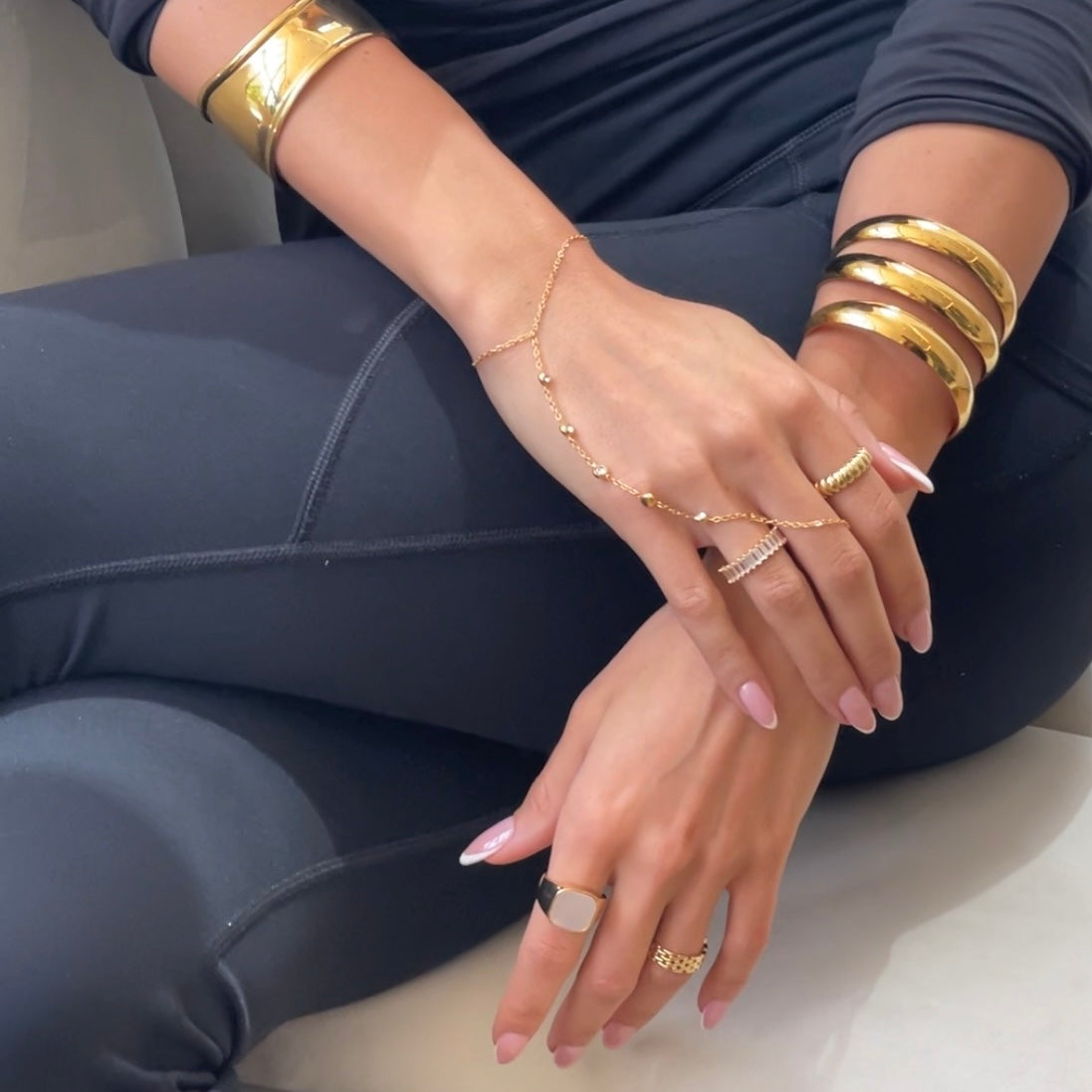 A close-up of a person wearing a gold chain hand bracelet with delicate bead details, paired with multiple gold bangles and rings, set against a dark gray leggings outfit.